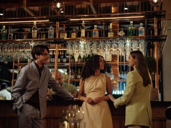 Three people smiling together at a wooden bar counter with a lit copper shelving display holding bottles and hanging glasses.