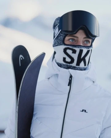 Close-up of a skier wearing a white ski jacket, patterned balaclava with ski text, black ski goggles, holding skis.