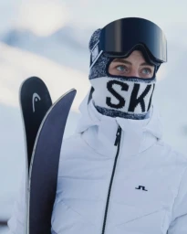 Close-up of a skier wearing a white ski jacket, patterned balaclava with ski text, black ski goggles, holding skis.
