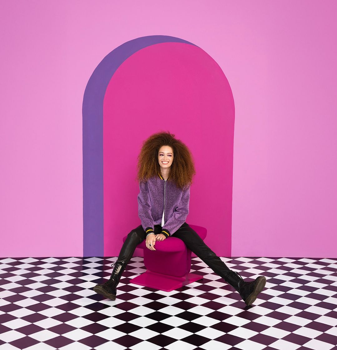 Young woman with afro sits with legs spread on pink seat in front of pink archway and black-and-white checkered floor, smiling.