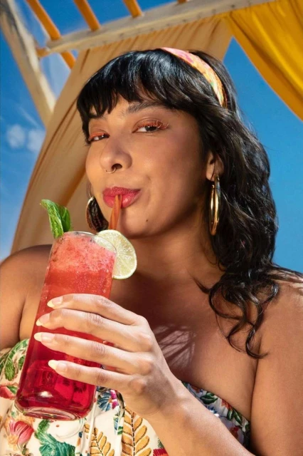 A woman with a patterned headband sips a red drink with a straw and a lime garnish against a blue sky and a tented awning.