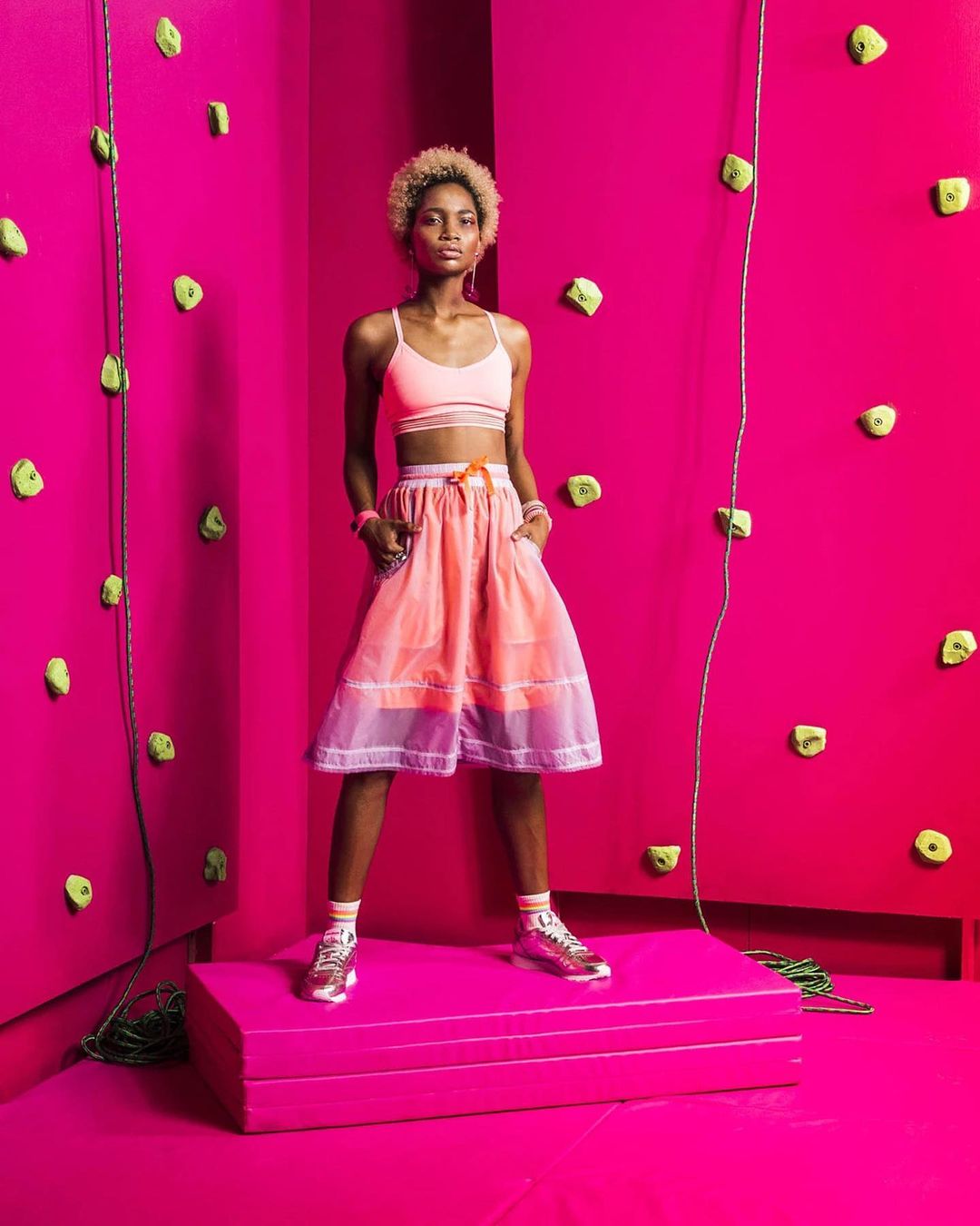 An athletic-looking young woman posing on a platform in a photo studio with a fuchsia pink background that resembles a climbing wall.