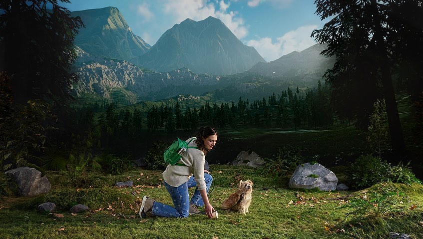 Woman kneeling on grass with her dog in front of a scenic mountain landscape backdrop, creating a nature-filled, staged photography scene.