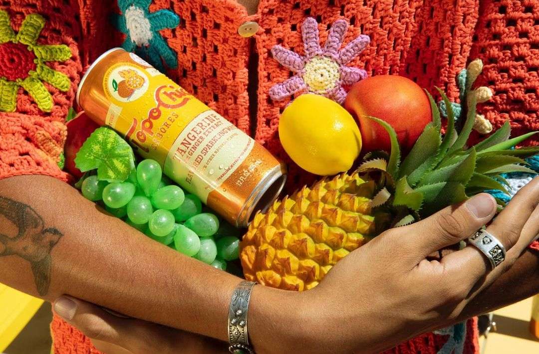 Person in crochet cardigan holds a colorful assortment of fruit and a Topo Chico tangerine sparkling water can in a bright, cheerful setting.