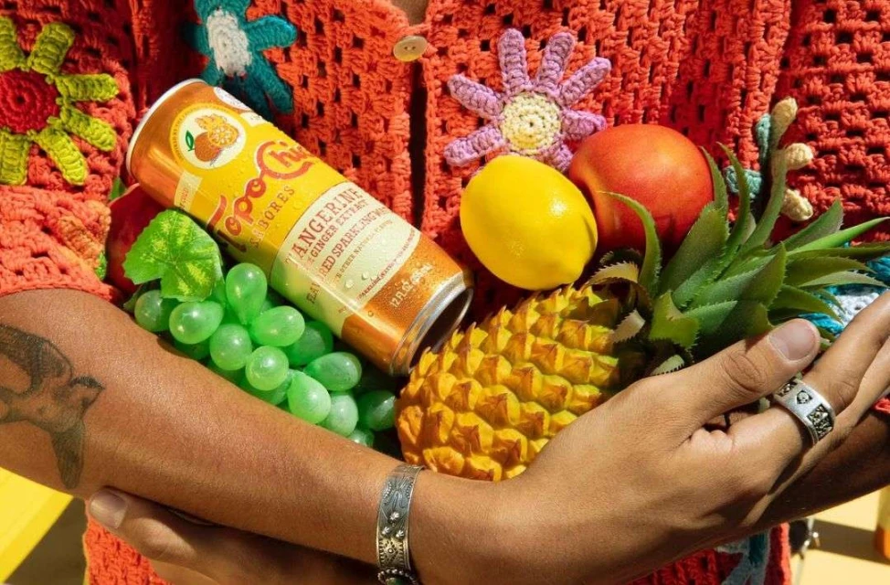 Person in crochet cardigan holds a colorful assortment of fruit and a Topo Chico tangerine sparkling water can in a bright, cheerful setting.