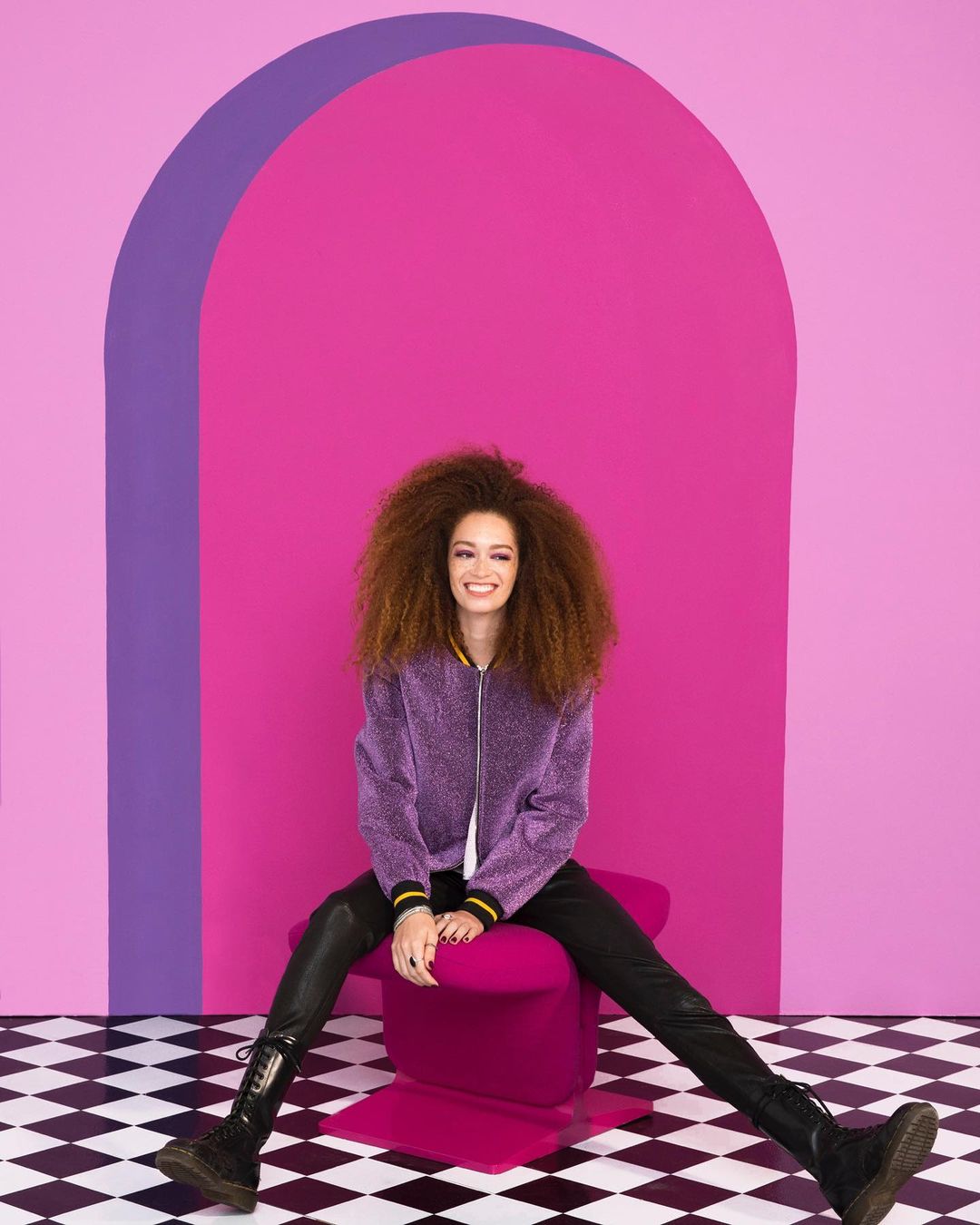 Smiling woman with afro sits on magenta chair against a pink and purple background. She wears a purple jacket, black pants and boots.