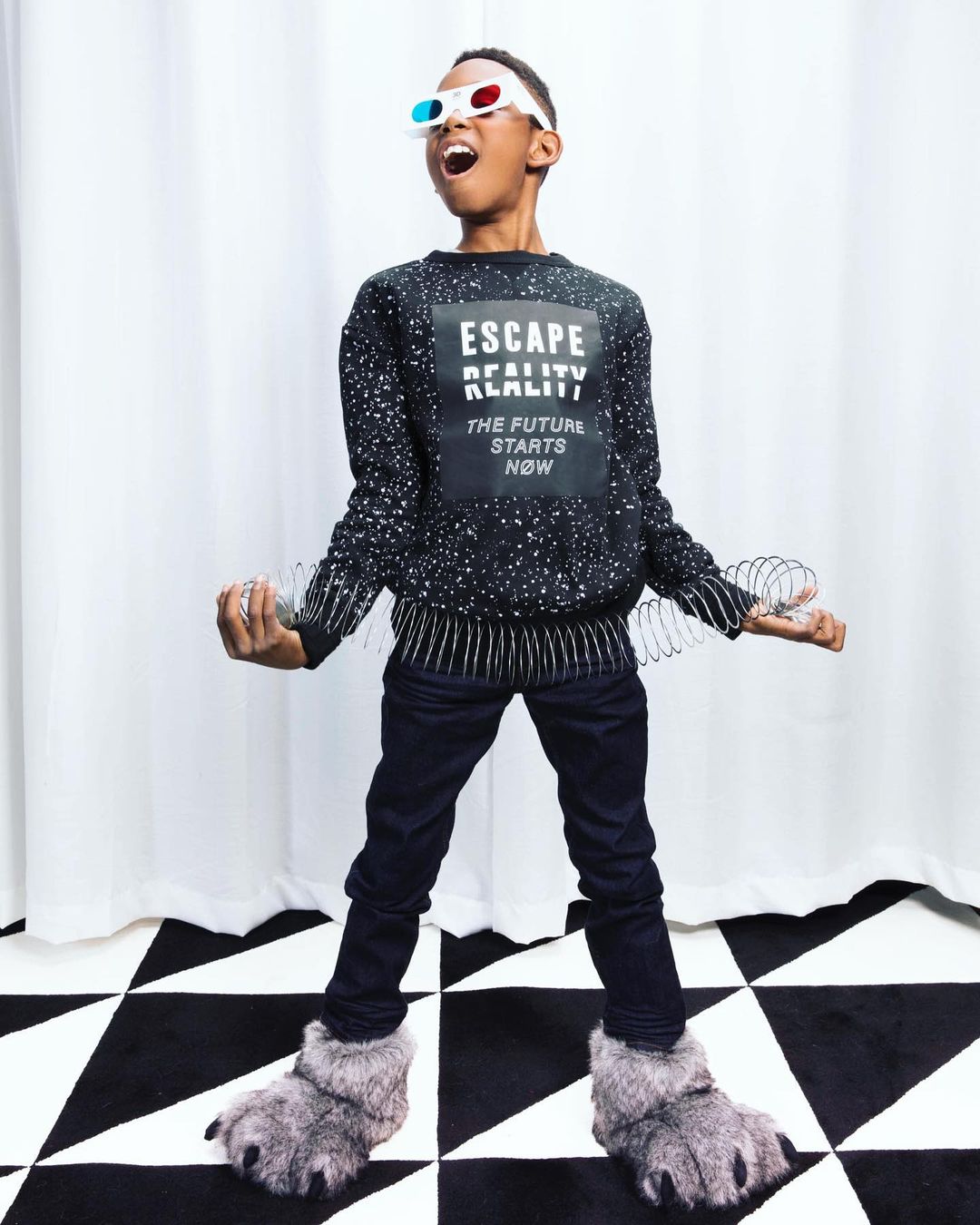 Child model in a speckled sweater, jeans, and furry slippers poses with a slinky on a black-and-white patterned floor.