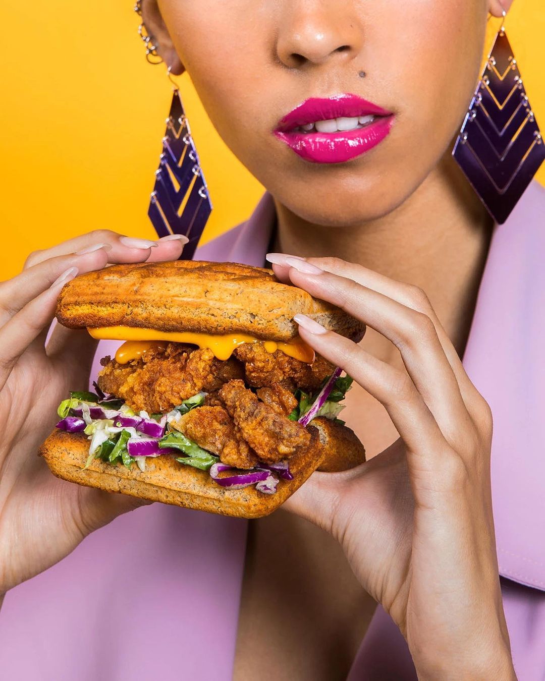 Woman in pink jacket holding a fried chicken sandwich with lettuce, tomato, and cheese against a yellow background in a studio shot.