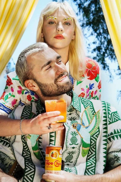 Two fair-skinned people pose for a photo: a blonde woman with decorative brows, and a bearded man holding a cocktail and Topo Chico.