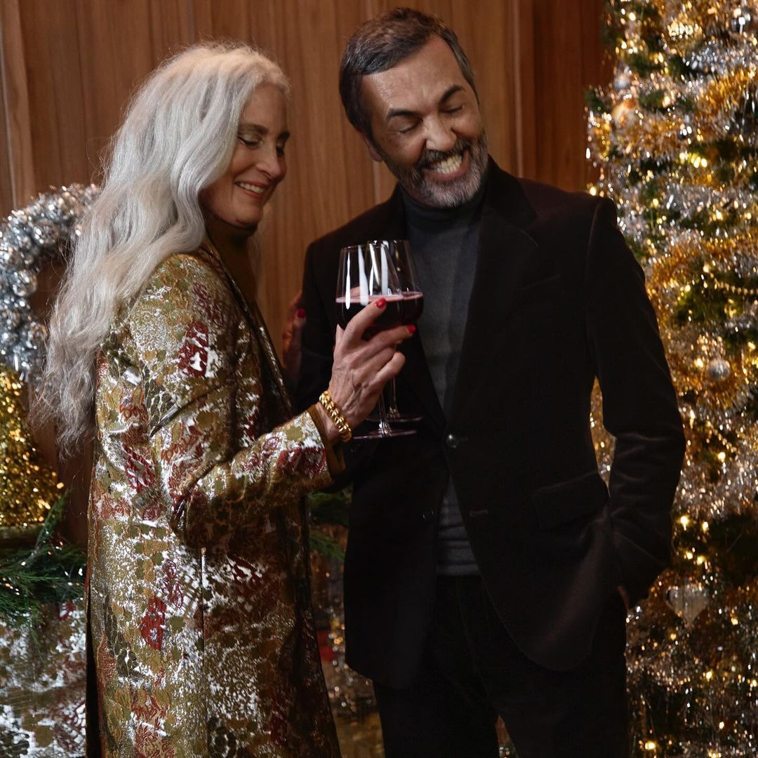 Mature couple smiling and holding a wine glass indoors with Christmas decor and wood-panelled walls, creating a cozy, festive atmosphere.