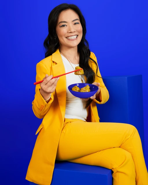 Smiling woman in a yellow suit is holding a bowl of dumplings with chopsticks against a solid blue background in a studio setting.