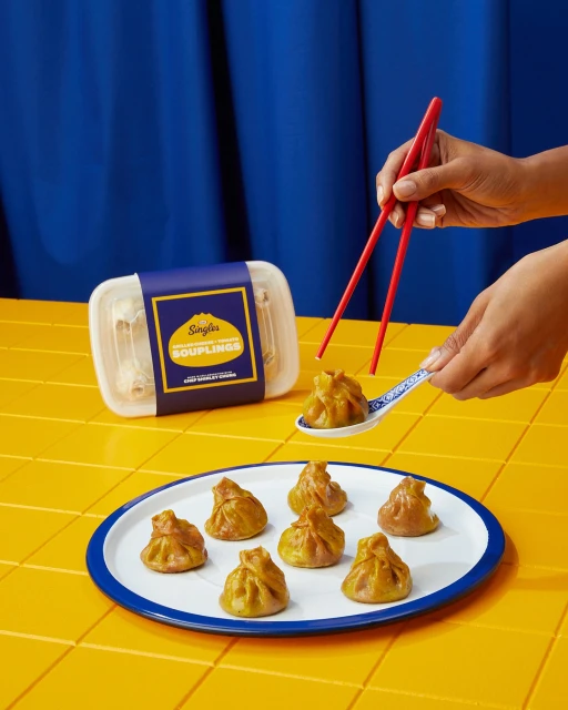 Close-up of a person using chopsticks to pick up a soup dumpling from a spoon, with more dumplings and packaging visible.