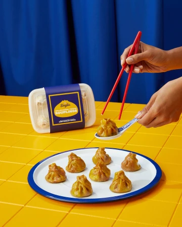 Close-up of a person using chopsticks to pick up a soup dumpling from a spoon, with more dumplings and packaging visible.