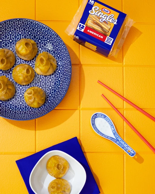 Overhead shot of dumplings on a blue patterned plate next to a package of Kraft Singles and red chopsticks on a yellow tile background.