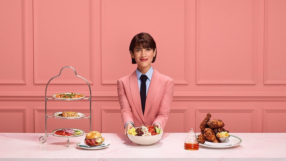A woman in a pink suit smiles while holding a salad bowl, with a platter of appetizers and a plate of fried chicken on a table.