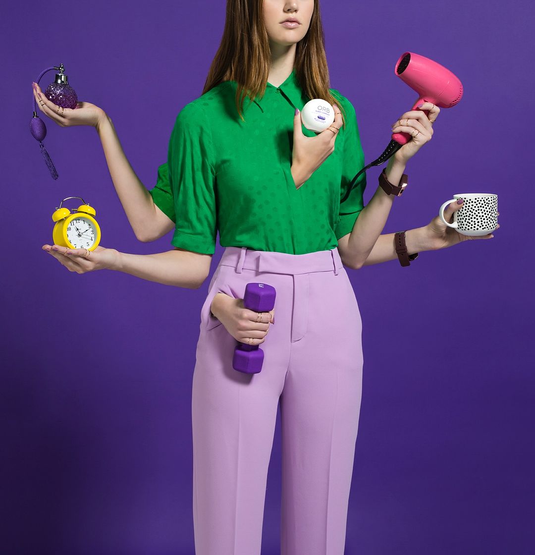 A woman with multiple arms holds an alarm clock, perfume bottle, face cream, dumbbell, hair dryer, and mug, set against a purple backdrop.