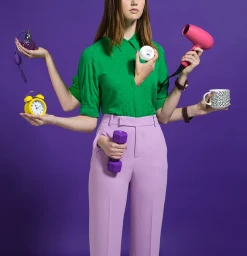 Woman wearing a green blouse and pink pants with multiple arms holding diverse objects against a solid purple background