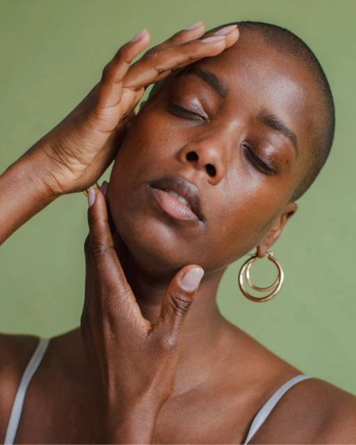 Close-up shot of a Black woman with shaved head and closed eyes, wearing hoop earrings, and posing with her hands near her face.
