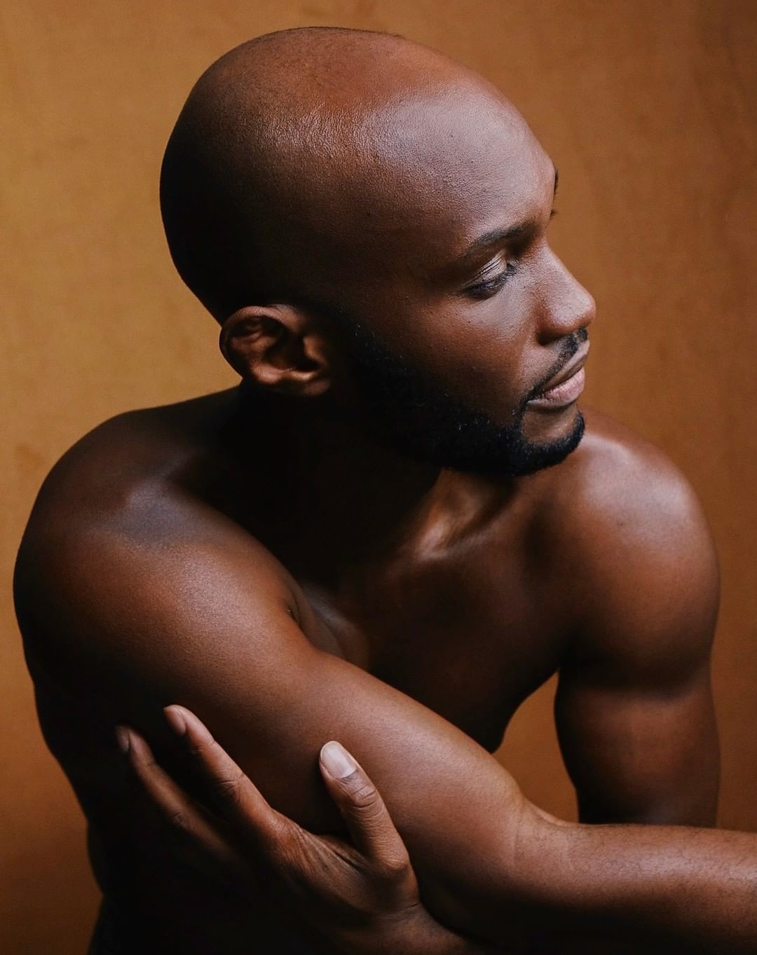 Close-up portrait of a shirtless, bald Black man against a tan background, looking off to the side with arms crossed.
