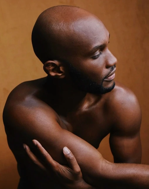 Close-up portrait of a shirtless, bald Black man against a tan background, looking off to the side with arms crossed.