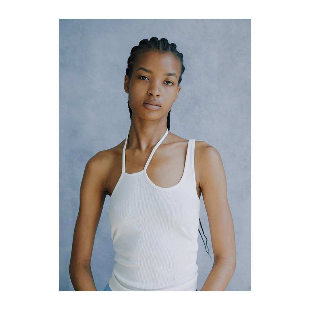 Close-up of a young adult female model with cornrow braids wearing a sleeveless white top against a light blue background.