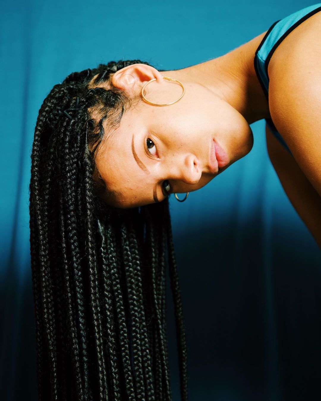 Portrait of a young adult female with long, braided black hair and gold hoop earrings, set against a blue background.