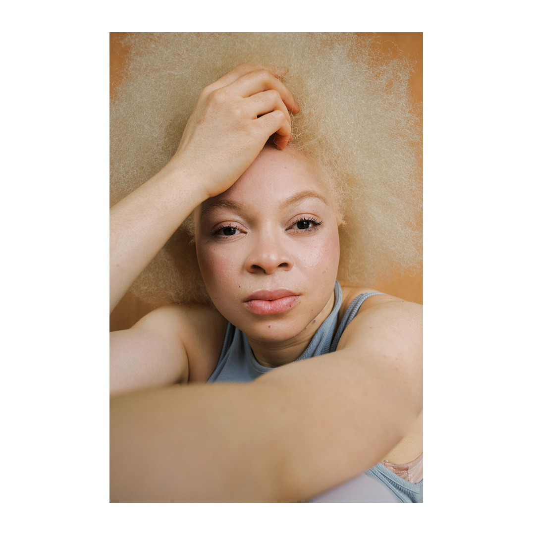 Close-up portrait of a woman with blonde hair, pale skin, and a neutral facial expression. She is wearing a grey top, posing indoors.