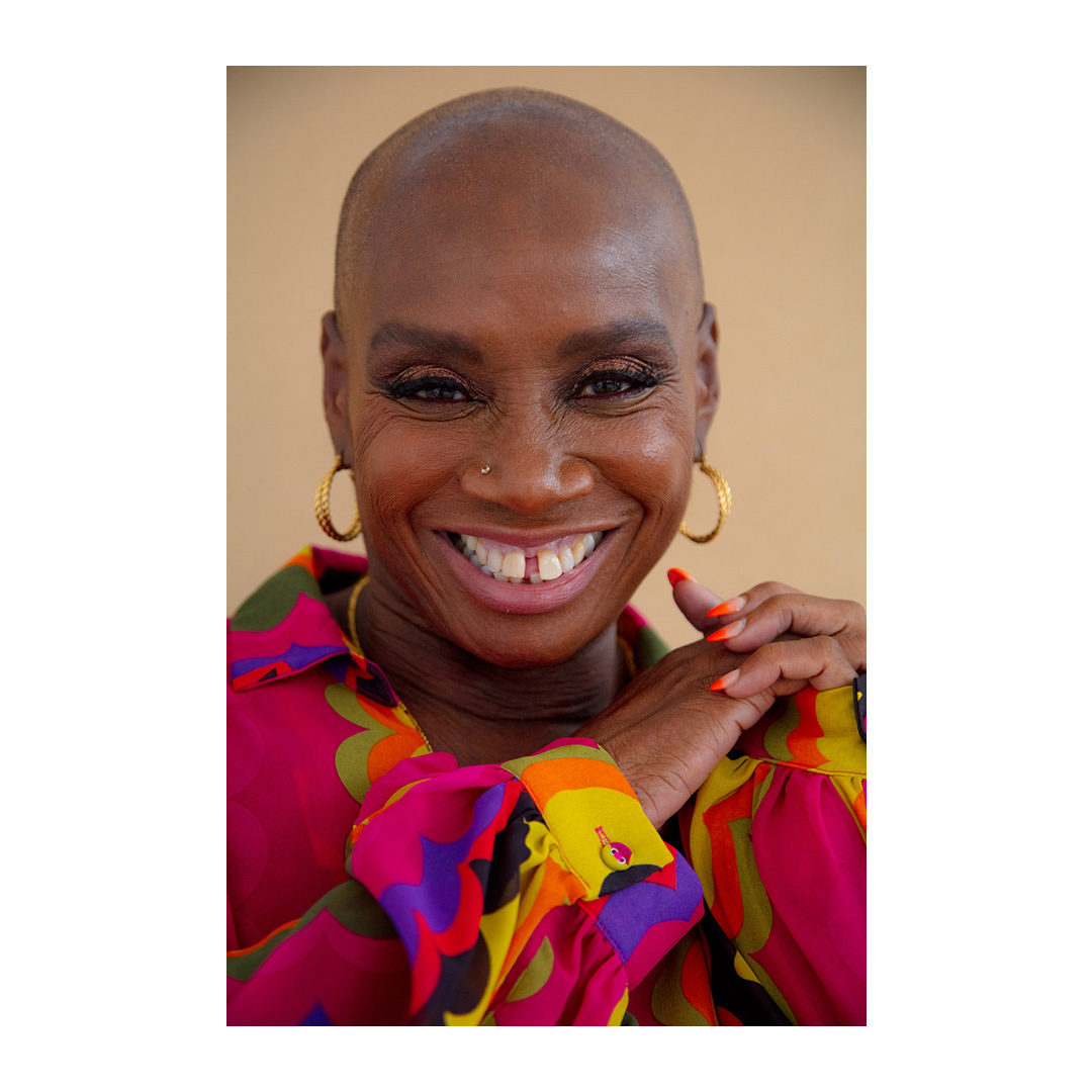 Close-up portrait of a smiling, bald Black woman with gold earrings and a colorful patterned shirt against a neutral background.