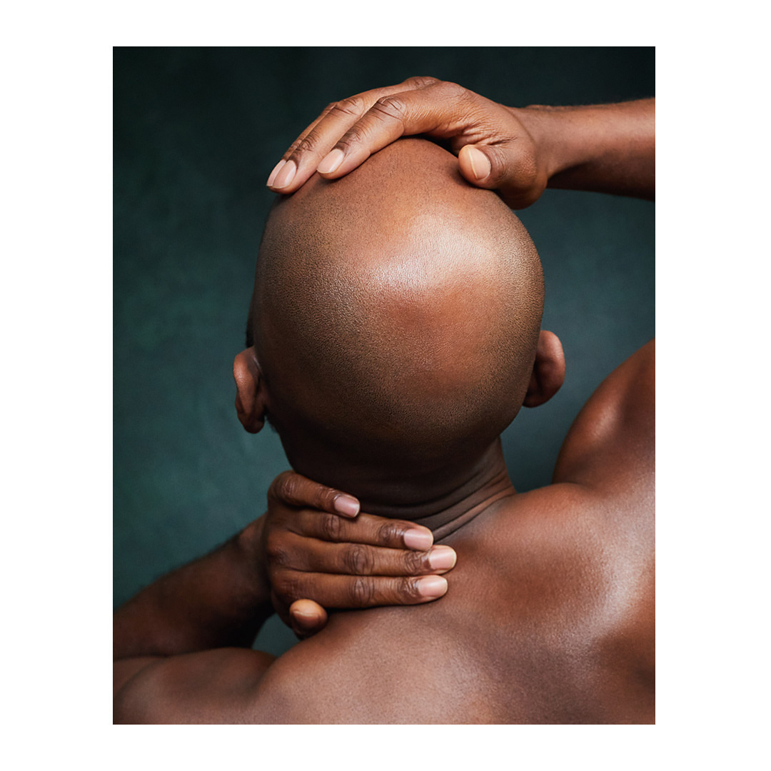 Close-up studio shot of a bald, dark-skinned adult male with his hands behind his neck, against a textured dark green background.