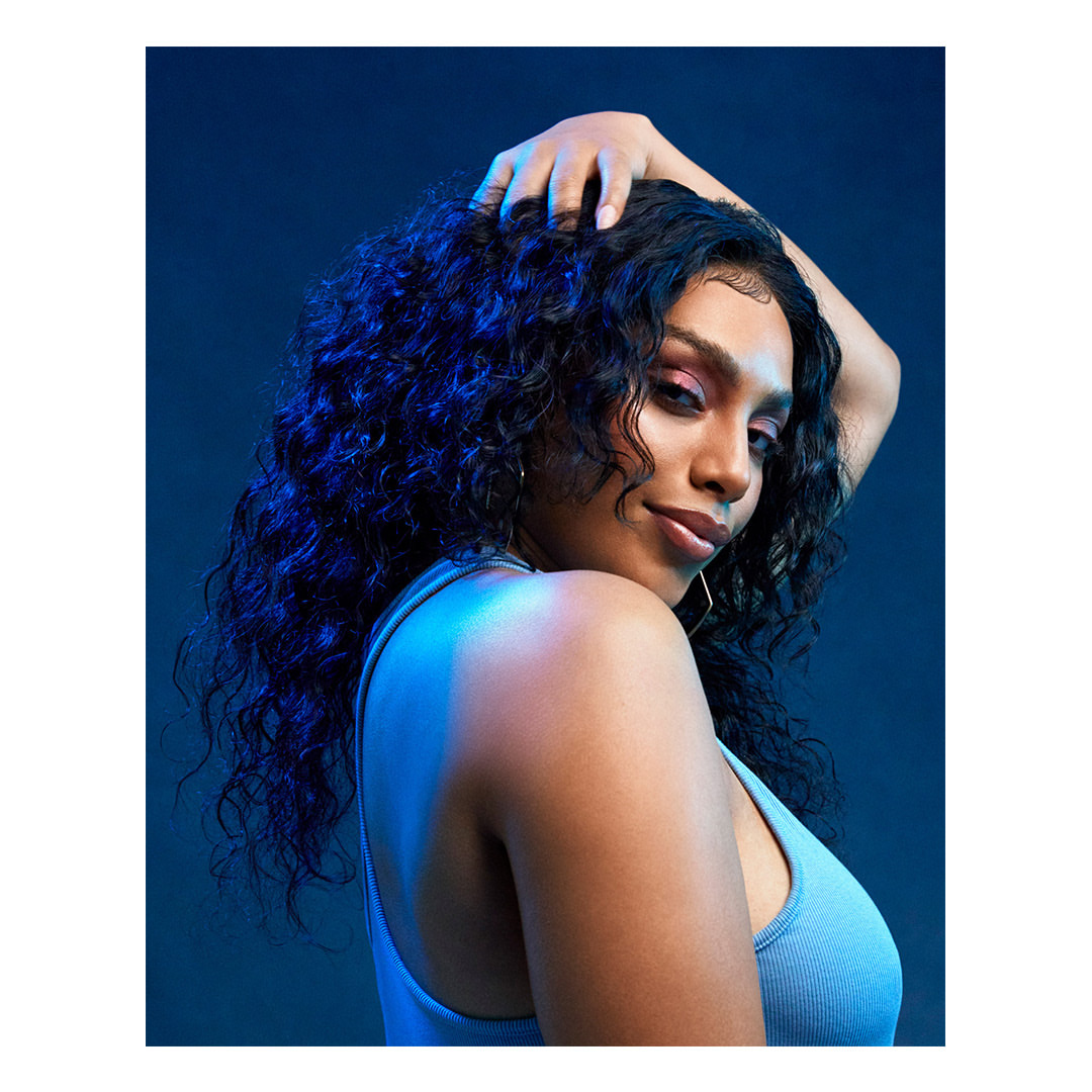 Portrait of a dark-skinned adult woman with curly hair, wearing a light blue tank top against a gradient blue background in a studio.