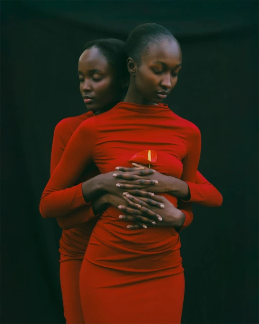 Two Black women in red dresses embrace, holding a red flower against a dark backdrop, creating a striking and intimate portrait.