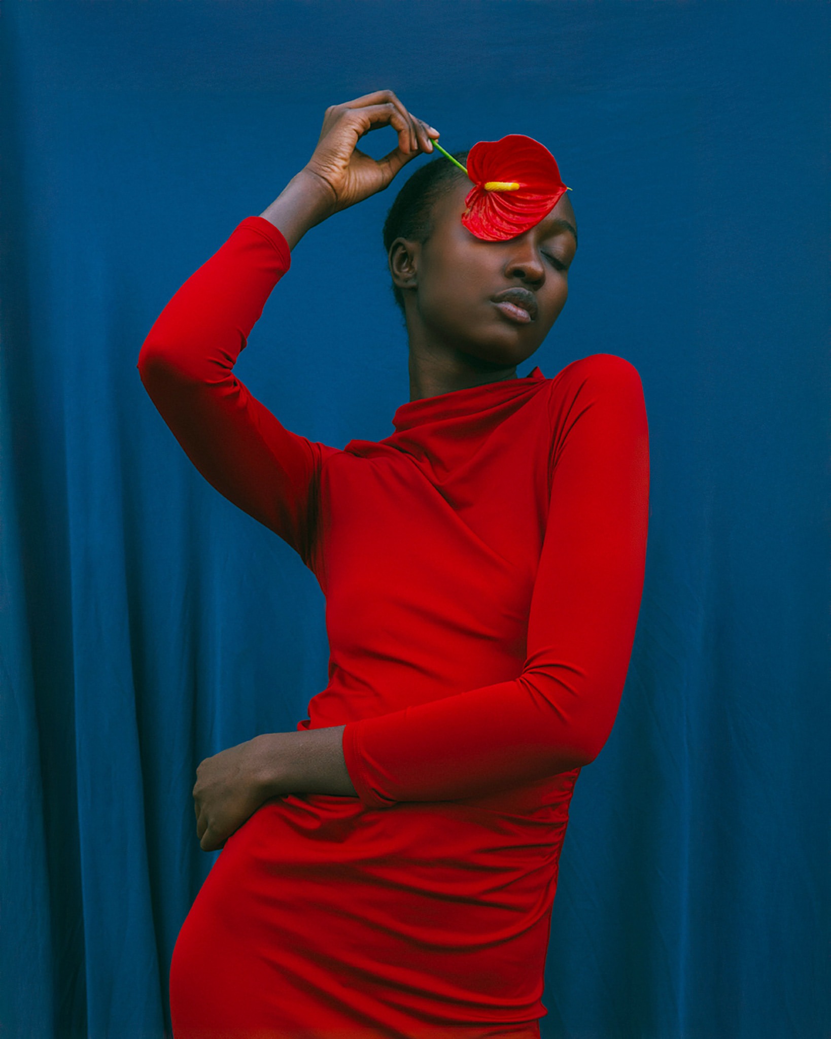 Elegant Black woman poses with a red anthurium flower in front of a blue curtain backdrop, wearing a matching red turtleneck dress.