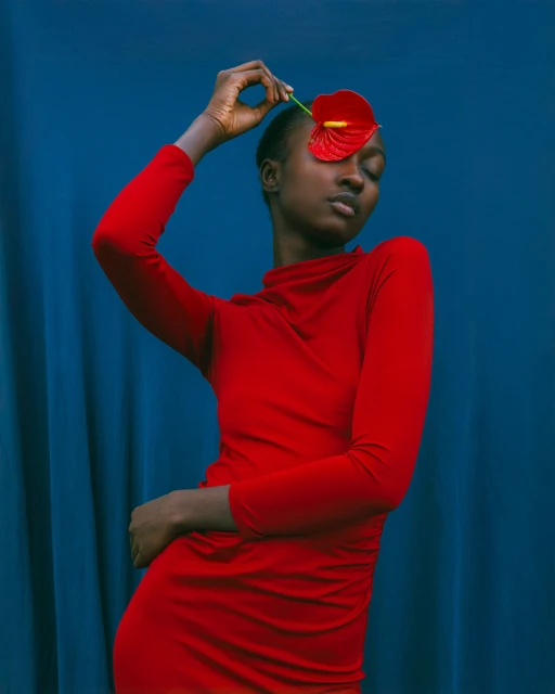 Elegant Black woman poses with a red anthurium flower in front of a blue curtain backdrop, wearing a matching red turtleneck dress.