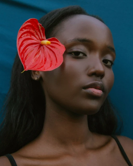 Close-up portrait of a young woman with dark hair and skin, wearing a black top, adorned with a red anthurium flower against blue backdrop.