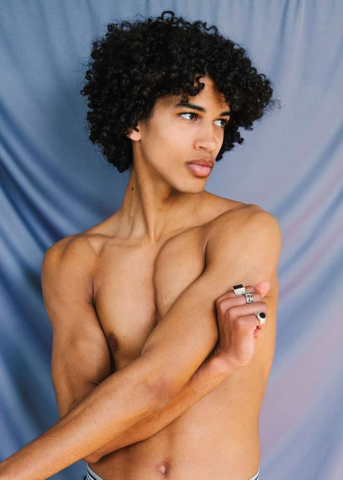 Shirtless young adult man with curly hair poses in a studio with a blue backdrop, showing his torso and arms, wearing rings.