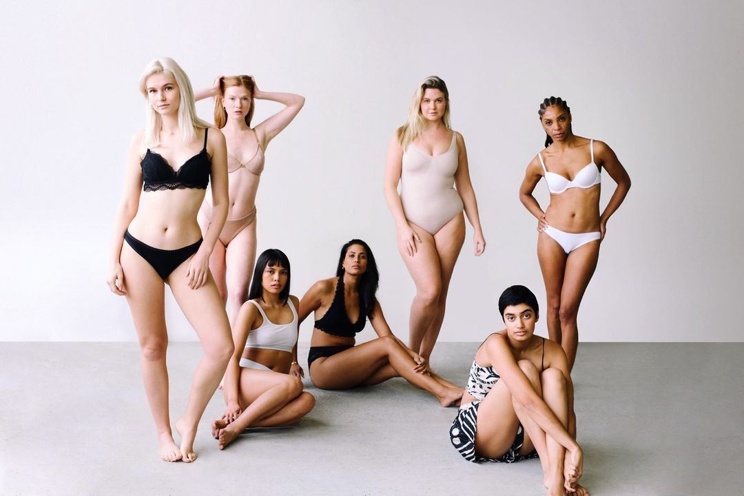 Seven women of diverse ethnicities and body types pose in lingerie against a white backdrop, promoting body positivity and inclusivity.