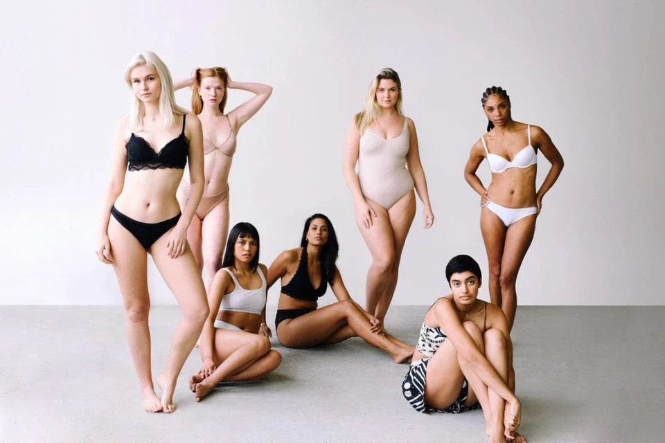 Seven women of diverse ethnicities and body types pose in lingerie against a white backdrop, promoting body positivity and inclusivity.