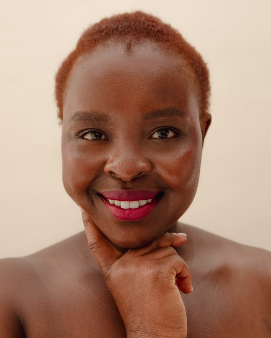 Close-up portrait of a smiling Black woman with short red hair, bright pink lipstick, and a hand gently touching her chin.