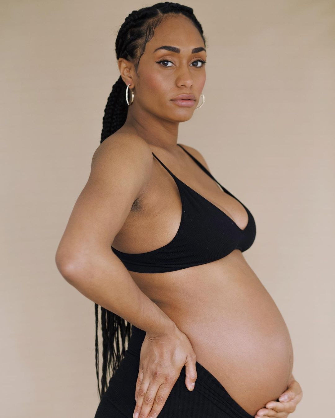 Side-view studio portrait of a pregnant adult woman with braids wearing black bra and pants against a neutral background.