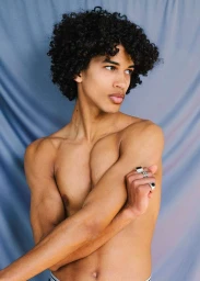 Shirtless young man with curly dark hair and silver rings posing against a draped blue fabric backdrop in a portrait by Krystal Neuvill.