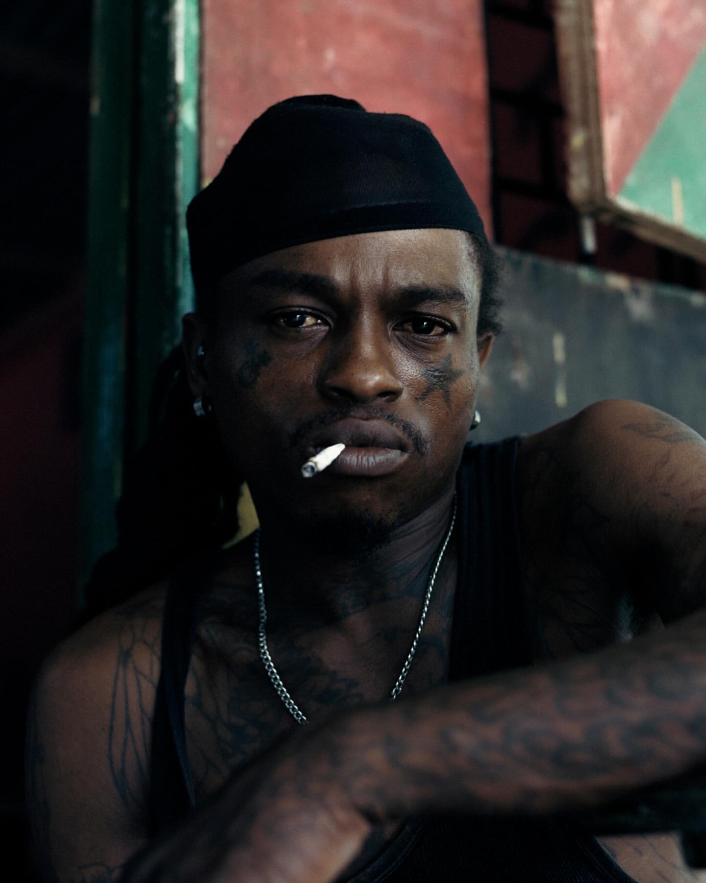 Portrait of a tattooed adult male with a cigarette, wearing a black beanie and silver chain, in a moody indoor setting with red and green.