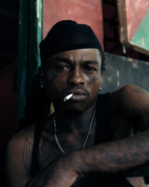 Portrait of a tattooed adult male with a cigarette, wearing a black beanie and silver chain, in a moody indoor setting with red and green.