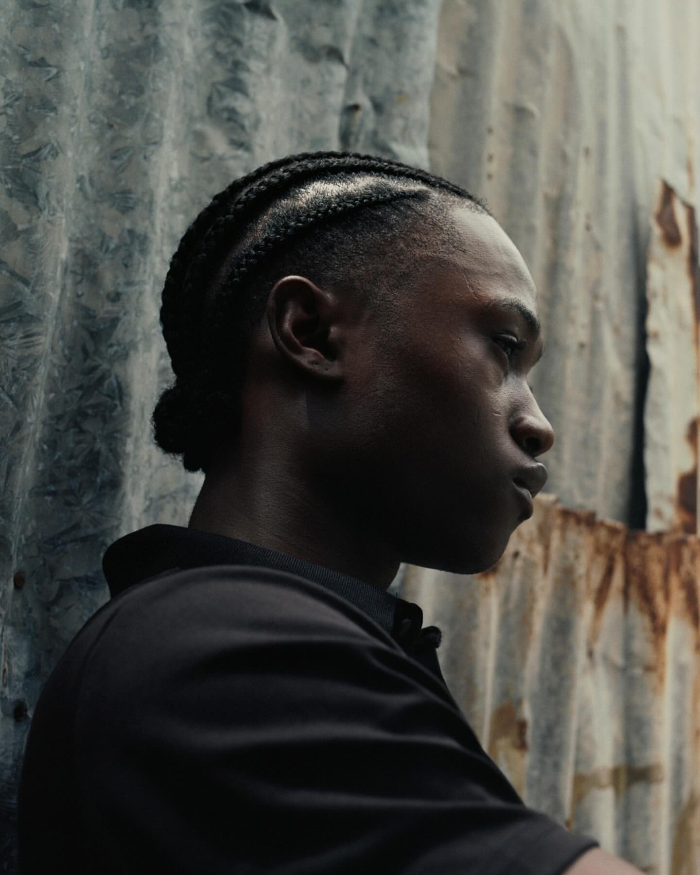 Profile portrait of an adult male with braided hair against a weathered corrugated metal wall in muted natural lighting.