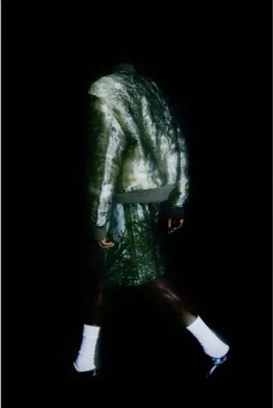 Figure in a green patterned bomber jacket and matching shorts walking against a solid black background.