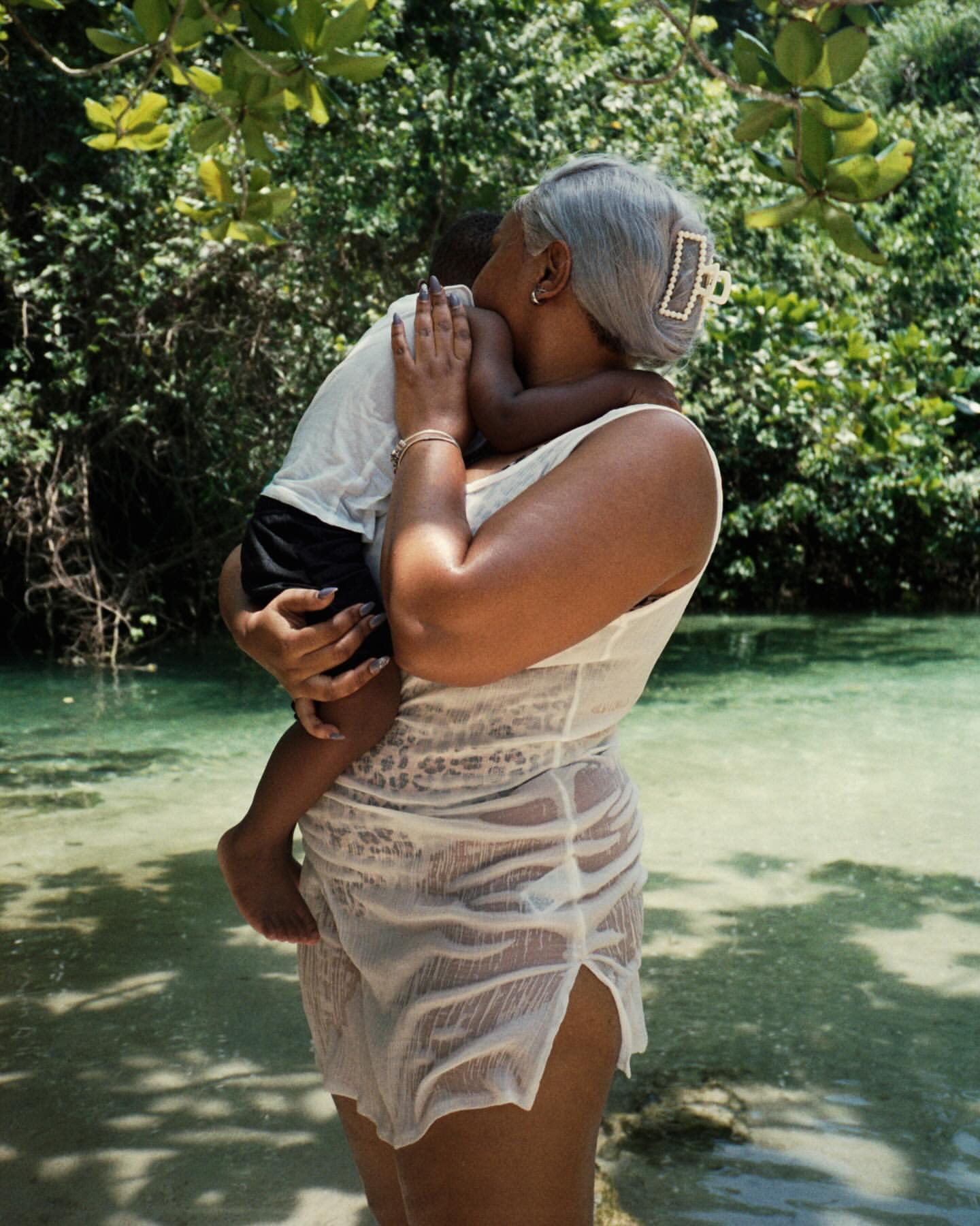 Woman standing in a body of water, holding a child. The background features lush trees and foliage, creating a natural, tranquil scene.