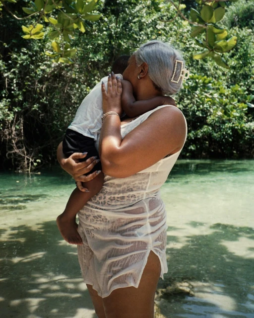 Woman standing in a body of water, holding a child. The background features lush trees and foliage, creating a natural, tranquil scene.