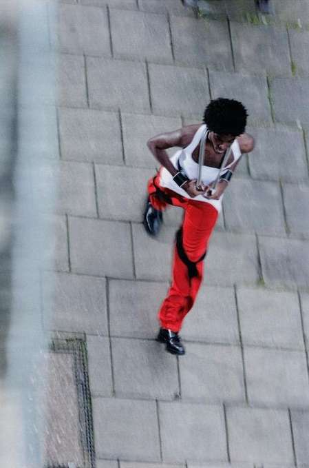 High-angle shot of an African-American woman in red pants and a white tank top dancing outside on a stone-paved area with chainlink fence.