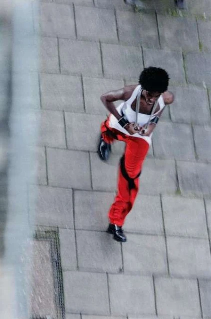 High-angle shot of an African-American woman in red pants and a white tank top dancing outside on a stone-paved area with chainlink fence.