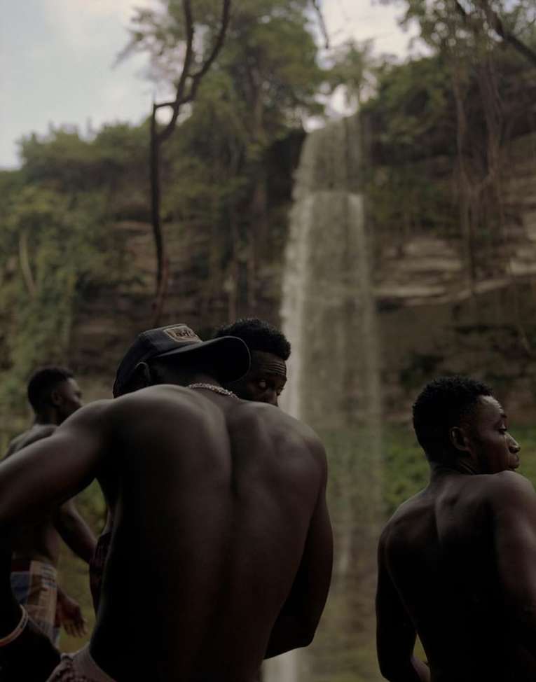 A group of men, some shirtless, gathered near a waterfall in a lush, green environment, captured with a focus on natural scenery.