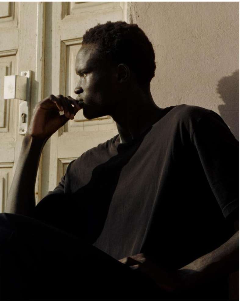 Portrait of a dark-skinned adult male wearing a casual t-shirt. He is seated with his hand raised to his mouth in a pensive pose.